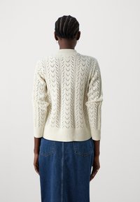 Häkelter cremefarbener Pullover mit einem gemusterten Design, gerippten Bündchen und Saum. Verfügt über einen runden Halsausschnitt und ein offenes Strickmuster. Getragen über einem Jeansrock.