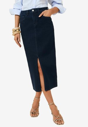 Falda midi de mezclilla oscura con una abertura frontal, que cuenta con una textura suave, cierre de botones y lazos para cinturón. Llevada con sandalias de tiras y pulseras de madera.