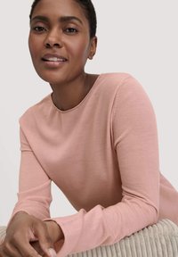 Sanftes rosa Langarmoberteil mit rundem Halsausschnitt und gerippter Textur, getragen mit Händen, die auf einer hellgrauen, plüschigen Oberfläche ruhen.