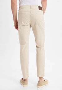 Beige Chinos mit schlanker Passform, leichtem Material, zwei Gesäßtaschen und einem braunen Lederpatch am Bund, kombiniert mit beigen Slippers.