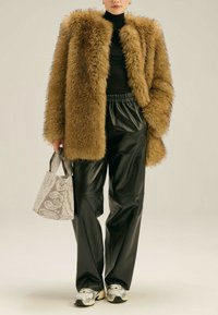 Brązowy sztuczny kożuch, czarny golf, błyszczące czarne skórzane spodnie, wzorzysty szary torebka i srebrne trampki, na neutralnym tle.