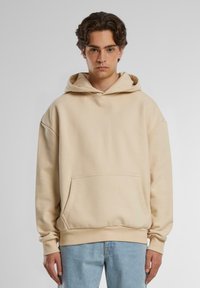 Beige hoodie with a drawstring hood, oversized fit, kangaroo pocket, ribbed cuffs, and hem. Made of soft fabric, suitable for casual wear.