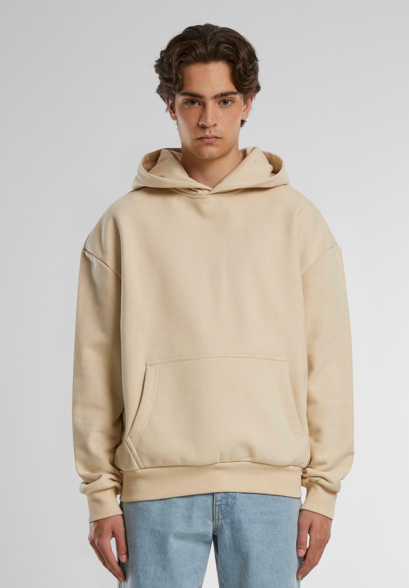 Beige hoodie with a drawstring hood, oversized fit, kangaroo pocket, ribbed cuffs, and hem. Made of soft fabric, suitable for casual wear.