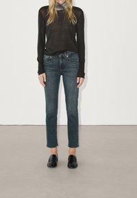 Woman wearing a sheer black long-sleeve top over a patterned collar shirt, dark blue straight-leg jeans, and black loafers standing indoors.