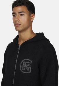 Młody mężczyzna w czarnej bluzie z zamkiem i szarym okrągłym logo na klatce piersiowej, zwrócony w lewo na jednolitym tle.