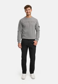 Smilende mann står iført en grå genser med teksten "INDICODE JEANS", svarte jeans og hvite joggesko mot en ensfarget hvit bakgrunn.