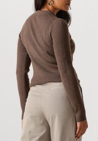 Frau trägt einen braunen gerippten Langarmpullover und beigen Hosen, von hinten und von der Seite gezeigt, mit goldenen Ohrhängern.