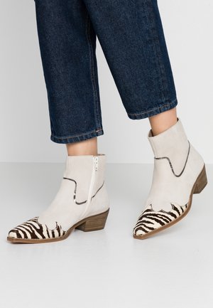 Personne portant des bottines écrues avec des bouts pointus imprimés zèbre et des talons en bois de hauteur moyenne, avec un jean court bleu foncé.