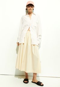 Helles rosa Hemd mit Knöpfen, cremefarbener, bodenlanger Rock, schwarze Riemensandalen und eine pinke Mütze. Einfaches Design, weiche Texturen und neutrale Farben.