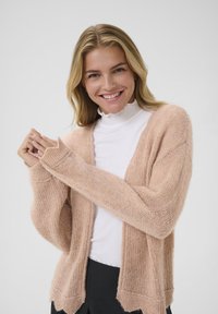 Cardigan de punto rosa claro con un acabado suave y texturizado, de frente abierto y un dobladillo sutilmente ondulado, usado sobre una blusa blanca de cuello alto.