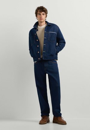 Joven de pie con chaqueta y pantalones vaqueros azul oscuro, suéter de punto color crema y zapatos de cuero marrón sobre un fondo liso y claro.