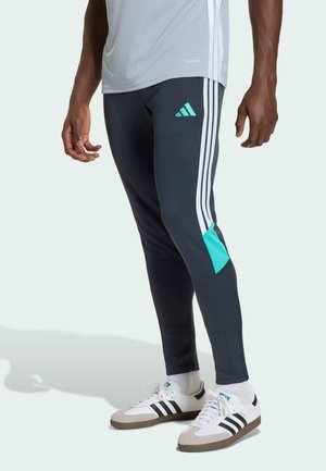 Mężczyzna ubrany w granatowe spodnie dresowe Adidas z białymi paskami i turkusowymi akcentami, białe buty sportowe Adidas i jasnoszarą koszulkę.
