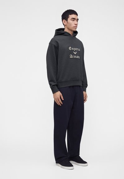 Νέος άνδρας φοράει σκούρα γκρι φούτερ Emporio Armani, μπλε παντελόνι και μαύρα αθλητικά παπούτσια, σε απλό φωτεινό φόντο.