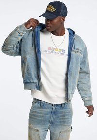 Giacca bomber in denim azzurro chiaro con polsini e orlo a coste. Indossata sopra una felpa bianca con logo multicolore. Jeans abbinati completano l'outfit.