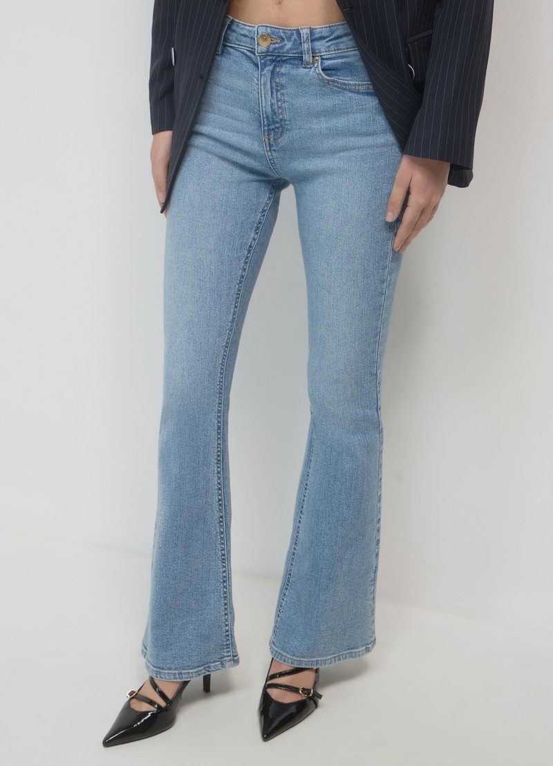 Jeans évasés bleu clair en denim avec une taille haute, présentant un lavage subtil et des coutures piquées, associés à des talons aiguilles noirs.