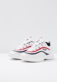 Zapatillas deportivas blancas con parte superior de malla y acentos en rojo y azul marino, un diseño curvado y una suela de goma texturizada. Logo mostrado en el lateral.