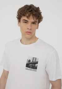 T-shirt in cotone bianco con stampa grafica in bianco e nero di un ponte e testo sulla parte sinistra del petto. Maniche corte e scollatura rotonda.