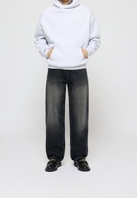 Sudadera gris con un bolsillo frontal, combinada con jeans negros de corte holgado y efecto desgastado, y zapatos negros con una cadena decorativa.