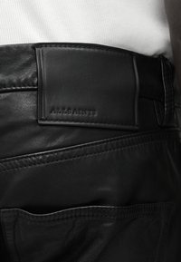 Μαύρο δερμάτινο παντελόνι με ραμμένες λεπτομέρειες και δερμάτινη ετικέτα με ανάγλυφο το "ALLSAINTS" στη μέση, φορεμένο με λευκό τοπ.