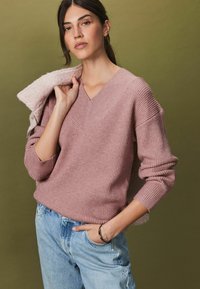 Harmaan vaaleanpunainen ribattu neule, jossa on V-pääntie ja väljät hiha, yhdistettynä vaaleansinisiin farkkuihin, joissa on hienovaraisia koristeluja. Pehmeä tekstuuri.