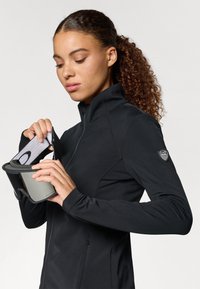 Jachetă sport neagră cu guler înalt și fermoar. Prezintă un logo pe mânecă și o persoană care ține ochelari de schi cu lentilă reflectorizantă.