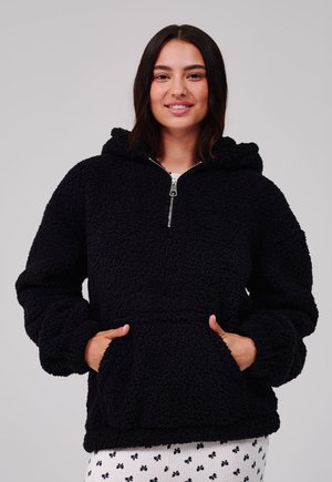Femme souriante portant un sweat à capuche en polaire noire avec fermeture éclair et poche kangourou, debout les mains dans les poches devant un fond uni.