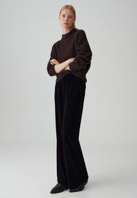 Bruine gebreide trui met een hoogneklintuur, gecombineerd met zwarte, wijd uitlopende corduroy broek en zwarte, puntige enkellaarsjes. Simpel ontwerp, minimale accenten.