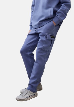 Person iført blå cargo sweatpants og matchende sweatshirt med hænderne i lommerne og grå sneakers, stående mod hvid baggrund.