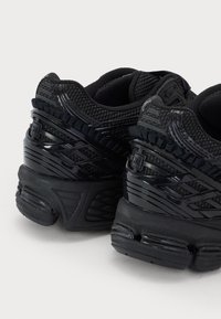Paire de chaussures de sport noires avec mesh texturé et détails en plastique brillant, vues de dos et orientées vers la droite, sur un fond blanc.
