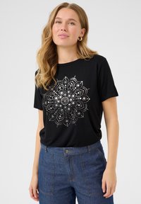 Sort t-shirt med et sølv mandala-design i midten, korte ærmer og en rund halsudskæring, parret med blå denim-bukser.