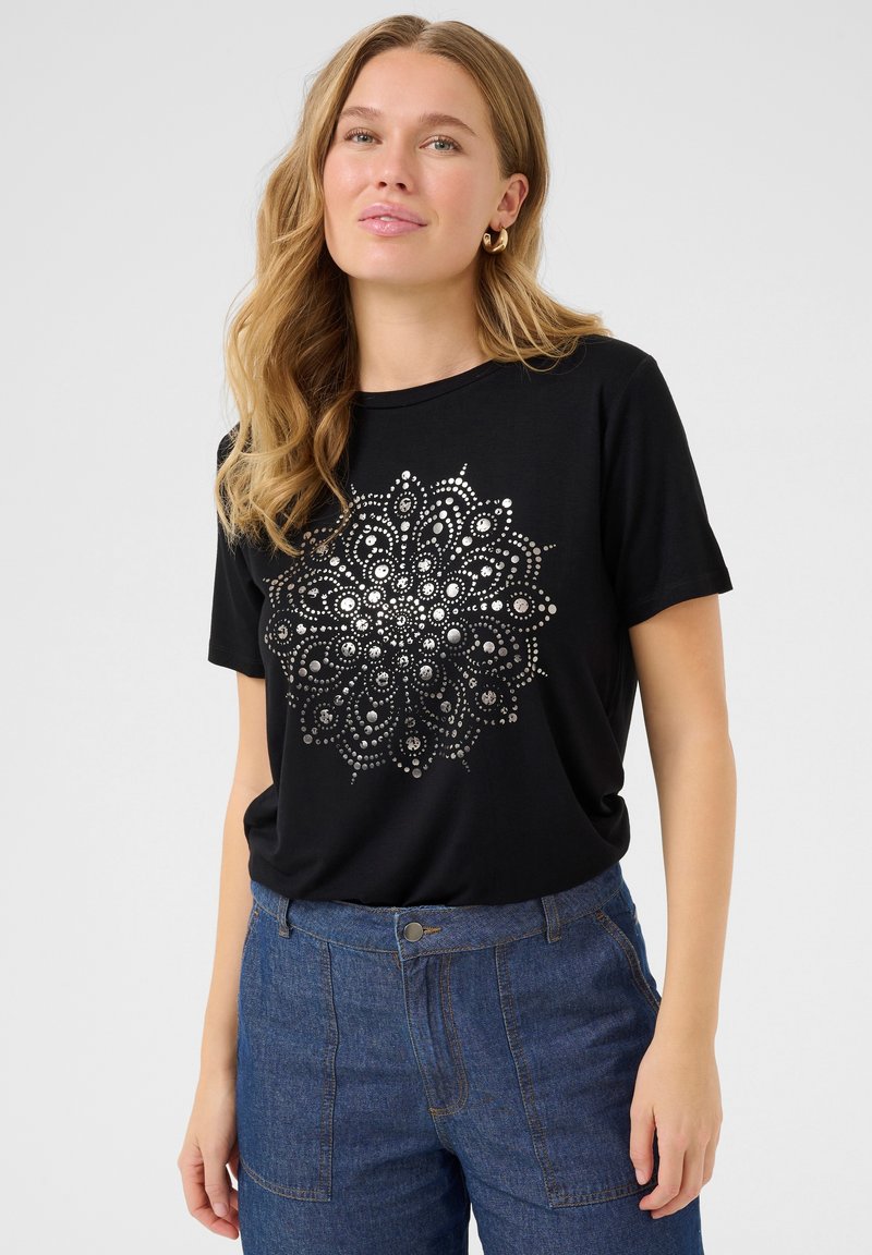 Sort t-shirt med et sølv mandala-design i midten, korte ærmer og en rund halsudskæring, parret med blå denim-bukser.