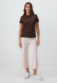 Braunes Baumwoll-T-Shirt mit kurzen Ärmeln, kombiniert mit hellbeigen weiten Hosen. Das Model trägt weiße Sneaker. Einfaches, lässiges Outfit mit minimalistischem Design.