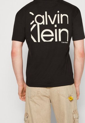 Black cotton t-shirt featuring large cream text "Calvin Klein" on the back. Short sleeves, crew neck, and relaxed fit with a side logo tag.
