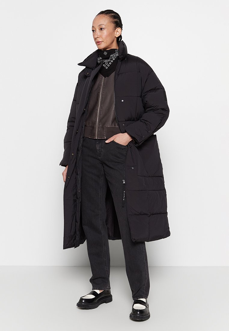 Frau trägt einen langen schwarzen Daunenmantel, eine Lederjacke, schwarze Jeans, schwarz-weiße Slipper und einen schwarz gemusterten Halstuch.