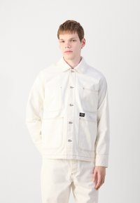 White denim jacket with a collar, front button closure, large pockets, contrast stitching, and a black brand label on the left side.