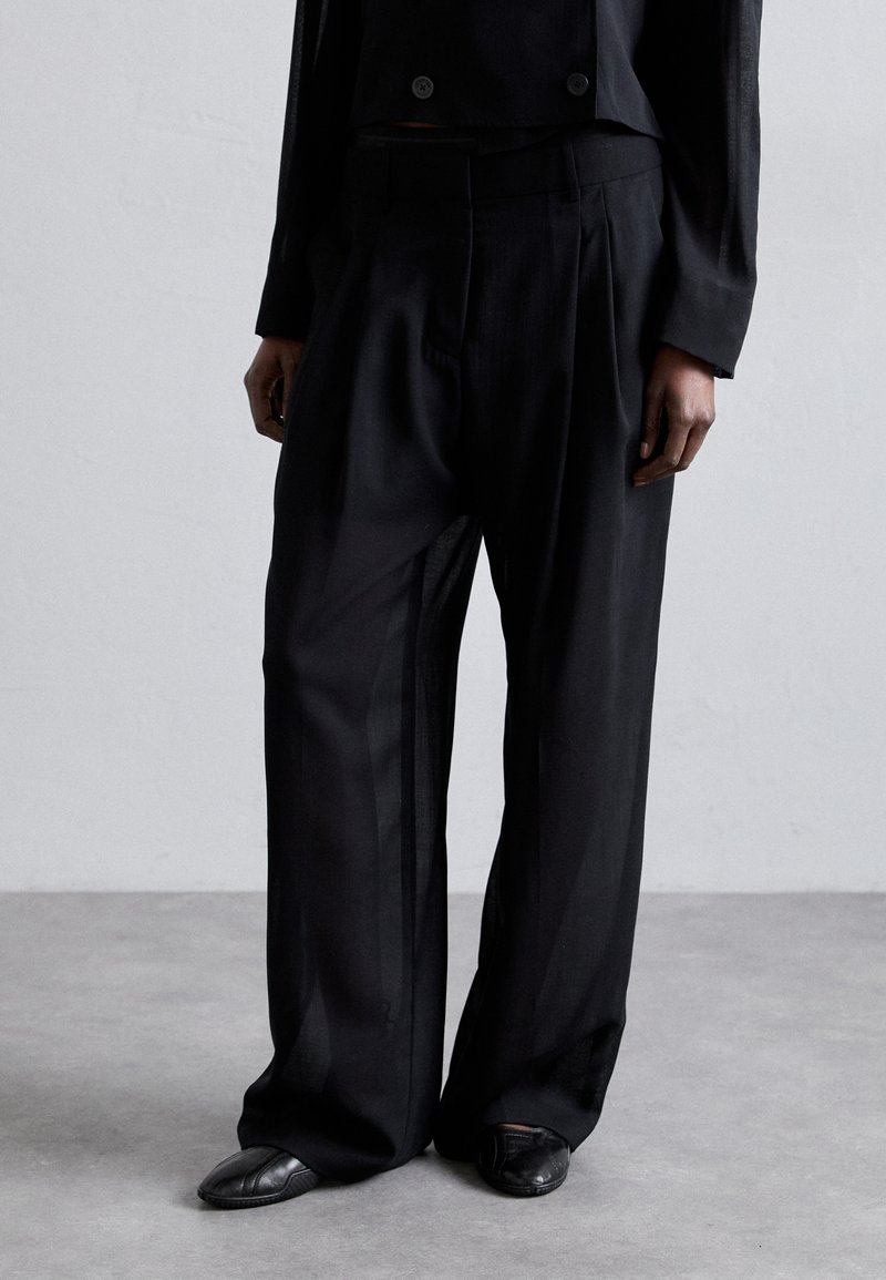 Črne široke hlače, izdelane iz lahkega materiala, z visokim pasom in blagimi gubami. Nosi se jih s preprostimi črnimi čevlji.