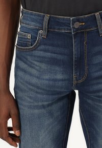 Jean en denim bleu foncé avec des coutures jaunes, comportant des poches avant et une fermeture à bouton à la taille. Texture lisse et design ajusté.