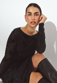 Melnas garām piedurknēm tērps ar ziedu rakstu, mīksts audums un pieguļoša silueta. Ceļa augstumā melni spīdīgi zābaki.