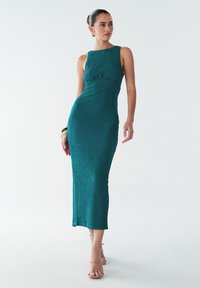 Žalsvai mėlyna, be rankovių, iki grindų siekianti suknelė iš tekstūruotos medžiagos, aptemptu liemeniu ir ryškia liemeniu. Dėvima su smėlio spalvos basutėmis su dirželiais.