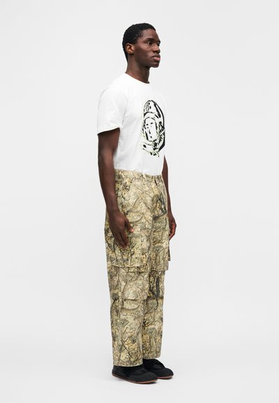 Λευκό μπλουζάκι με μαύρο και πράσινο γραφικό, συνδυασμένο με παντελόνια καμουφλάζ τύπου cargo και μαύρα αθλητικά παπούτσια. Στενή εφαρμογή και επίπεδη υφή.