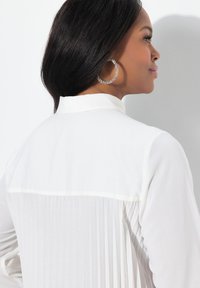 Witte blouse met geplooide achterkant, met een kraag. Model draagt grote ronde oorbellen, haar los gestyled. Soepele stoftextuur.