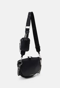 Zwarte schoudertas met een afneembare tas, brede strap met reliëftekst, zilveren kettingdetails en het "Steve Madden" logo aan de voorkant.