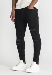 Čierne džínsy skinny strihu majú roztrhnuté detaily s rozstrapkanými kolenami. Sú skombinované s bielymi teniskami. Materiál vyzerá jemne a elasticky.