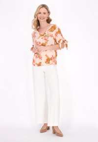 Blusa floreale con sfondo crema, pattern arancioni e verdi, e maniche legate, abbinata a pantaloni bianchi a gamba larga e tacchi beige.