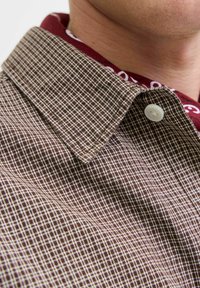 Brown checkered shirt with a white pattern, featuring a buttoned collar and a contrasting burgundy inner collar. Textured fabric detail visible.