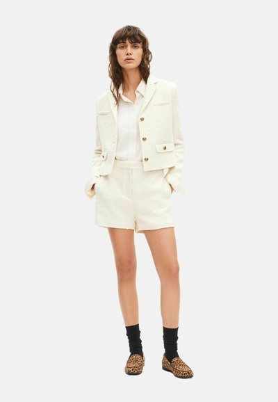 Femme portant un ensemble blazer et short texturé de couleur blanc cassé, blouse blanche, chaussettes noires et mocassins à imprimé léopard, debout les mains dans les poches.