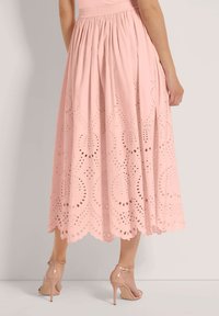 Rosa, baumwollener Midilänge-Rock mit gestickten Cutout-Mustern und gewelltem Saum. Kombiniert mit transparenten High-Heel-Sandalen.
