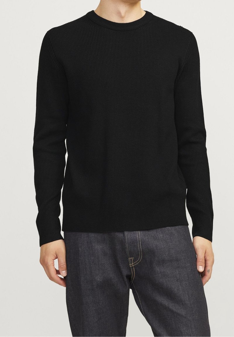 Homme portant un pull à manches longues en maille côtelée noire et un jean denim foncé, debout contre un fond clair uni.