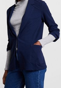 Blazer bleu marine en tissu doux avec une poche avant unique, des revers crantés et des manches longues, porté sur un col roulé gris.