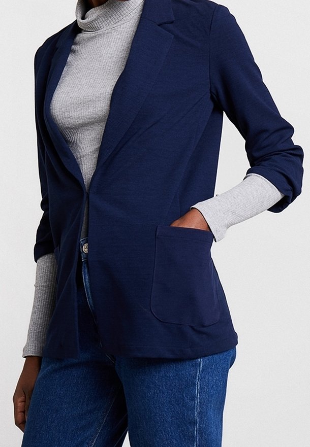 Blazer bleu marine en tissu doux avec une poche avant unique, des revers crantés et des manches longues, porté sur un col roulé gris.